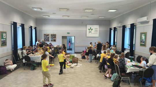 grupa uczniów siedzących przy stolikach, wykonują prace plastyczne, w tle sala widowiskowa
