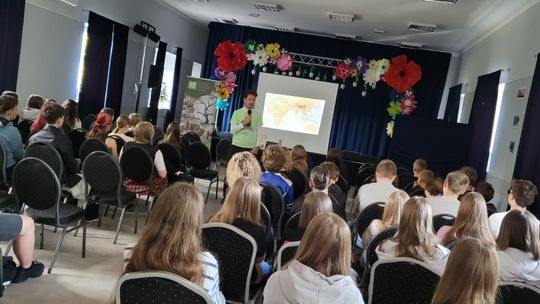 Prelegent prowadzi spotkanie dla młodzieży w sali konferencyjnej. Uczestnicy siedzą na krzesłach i słuchają wystąpienia. Za prelegentem widoczny jest ekran z prezentacją oraz dekoracja sceny z dużymi papierowymi kwiatami.