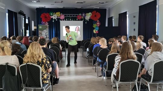 Prelegent przemawia do licznej grupy młodzieży zgromadzonej w sali konferencyjnej. W tle znajduje się ekran z prezentacją, a scena jest ozdobiona kolorowymi papierowymi kwiatami.
