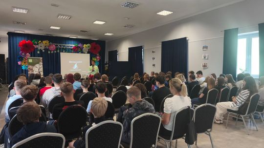 Prelegent przemawia do licznej grupy młodzieży zgromadzonej w sali konferencyjnej. W tle znajduje się ekran z prezentacją, a scena jest ozdobiona kolorowymi papierowymi kwiatami.