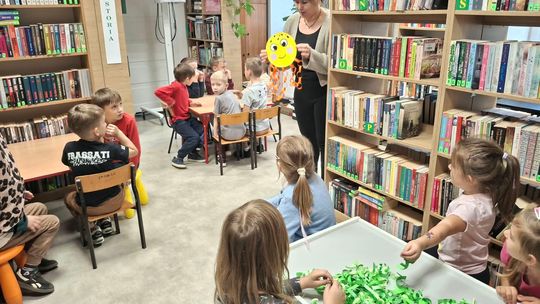 Dzieci siedzą przy stolikach i słuchają poleceń bibliotekarki na temat pracy plastycznej, którą mają wykonać. Bibliotekarka pokazuje im przygotowanę ośmiornicę.